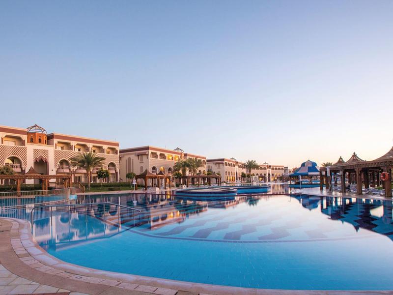 Velký prázdný venkovní bazén v resortu s palmami a hotelovými budovami při východu slunce.