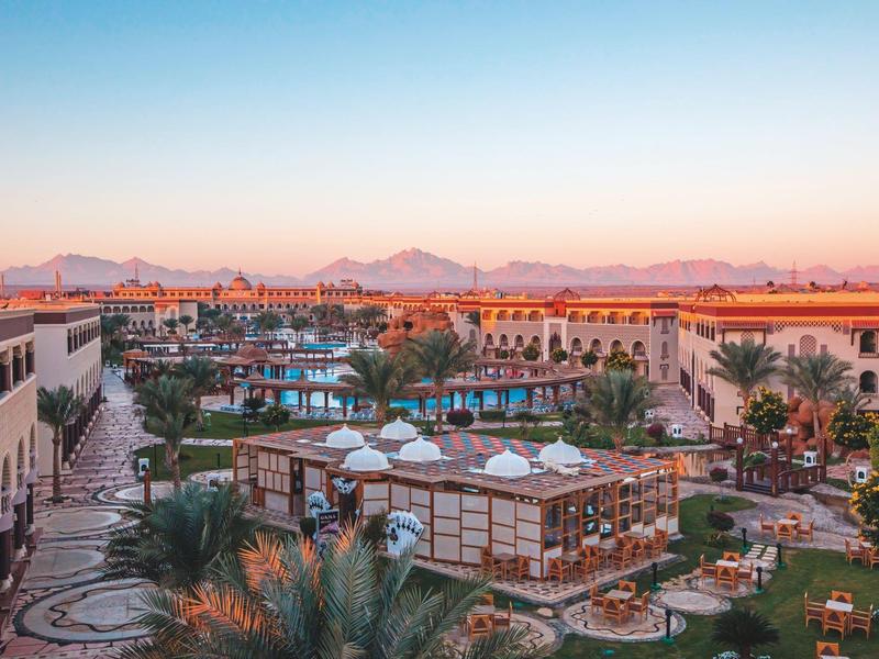 Resort s bazénem, palmami a horami v pozadí při západu slunce.