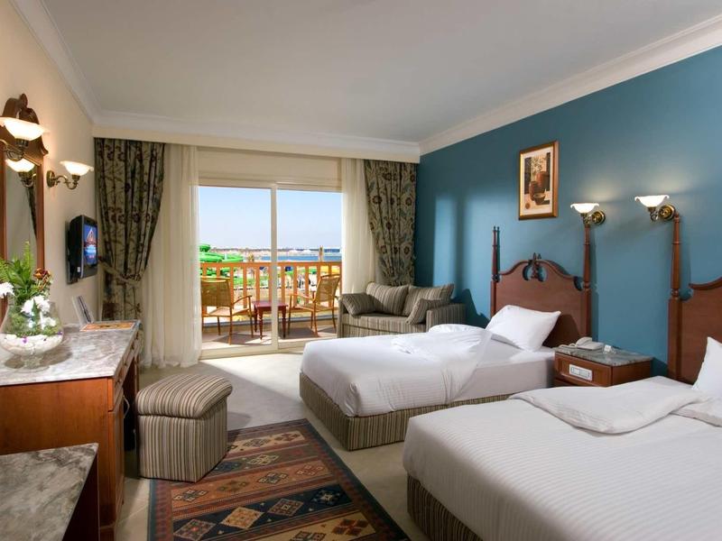 Hotelový pokoj se dvěma postelemi, posezením a balkonem s výhledem na zeleň.