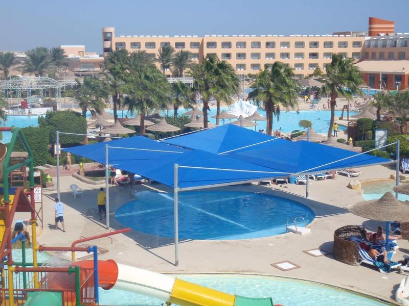 Hotelový bazén se slunečními plachtami, lehátky a palmami pod jasnou oblohou.