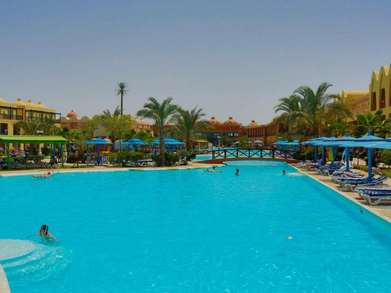 Velký hotelový bazén s lehátky a slunečníky, obklopený palmami a budovami.