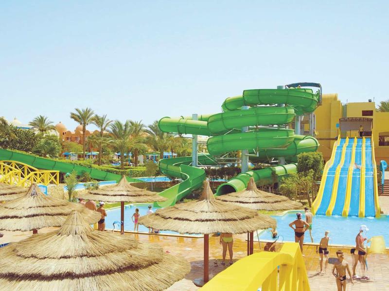 Vodní park s velkými zelenými a modrými skluzavkami, slunečníky a lidmi.