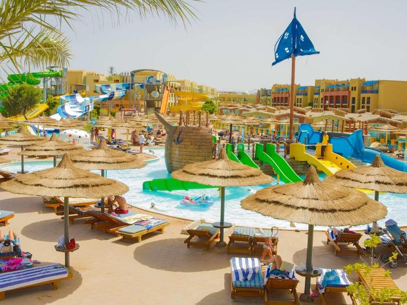Plážová oblast resortu s slunečníky, lehátky a vodními skluzavkami za slunečného počasí.