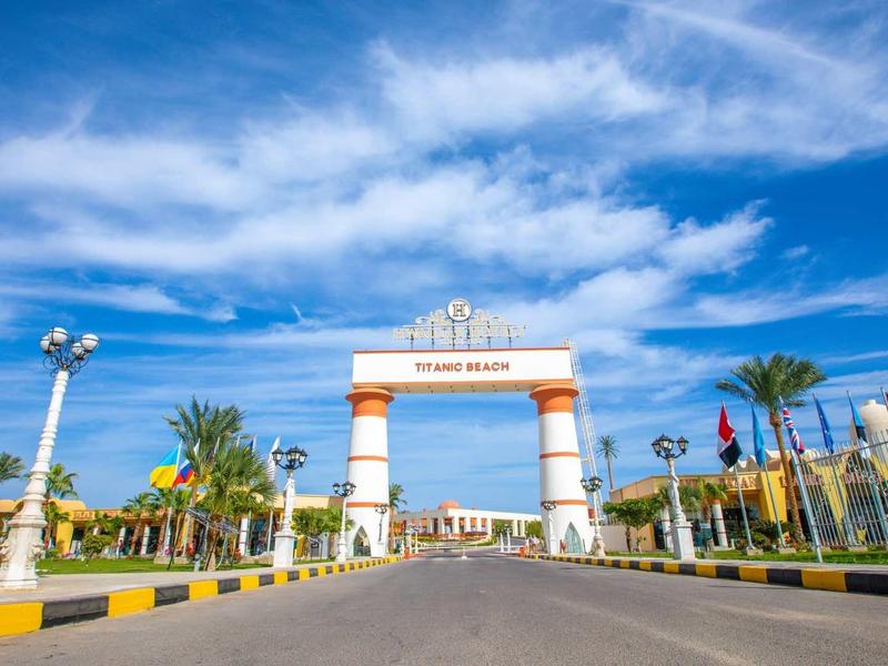 Vstupní brána hotelu s bledě modrou oblohou a palmami podél široké silnice.