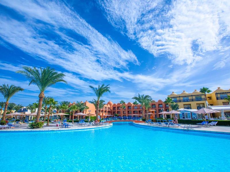 Velký modrý bazén s palmami a barevnými hotelovými budovami pod zamračenou modrou oblohou.