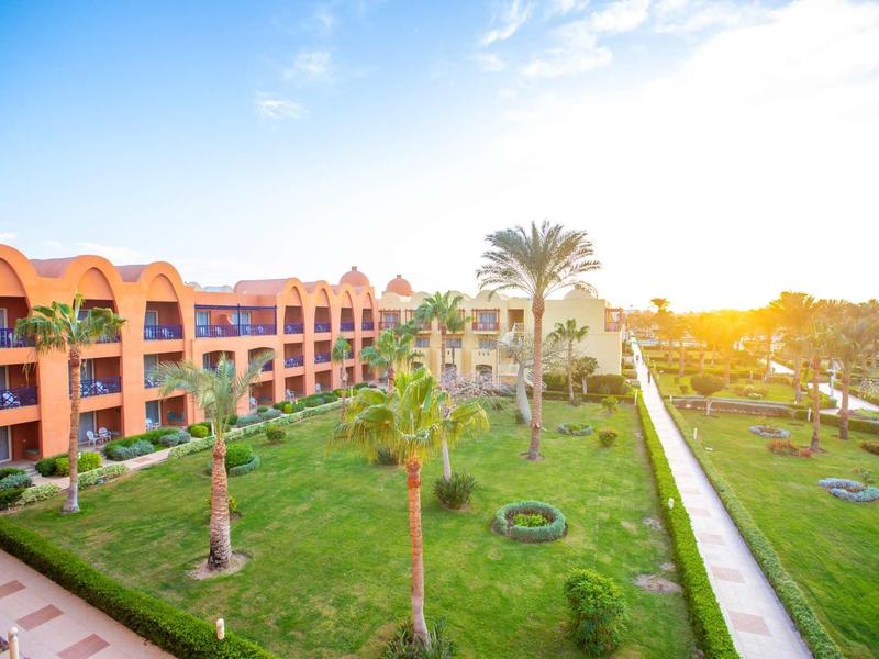 Hotelový komplex se zeleným trávníkem, palmami a budovami na slunci.