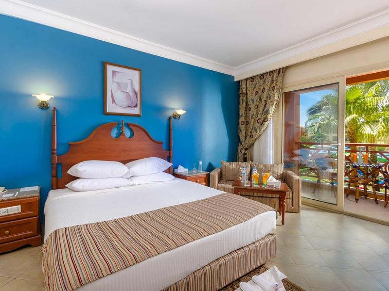 Světle zařízený hotelový pokoj se dvěma lůžky, posezením a balkonem s výhledem na palmy.