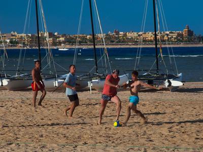 Cuatro hombres juegan fútbol playa en la arena con barcos de vela al fondo bajo cielo despejado.