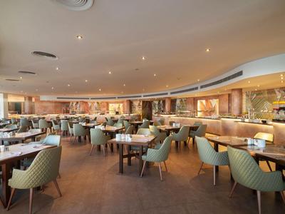 Un restaurante moderno y espacioso con muchas mesas y sillas acolchonadas en tonos cálidos.