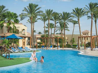 Gran piscina con tumbonas y palmeras en un resort hotelero bajo un clima soleado.