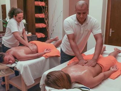 Dva hosté leží na masážních stolech a dostávají masáže zad od dvou masérů.