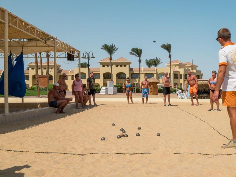 Lidé hrají pétanque na slunném písčitém hřišti s hotely v pozadí.