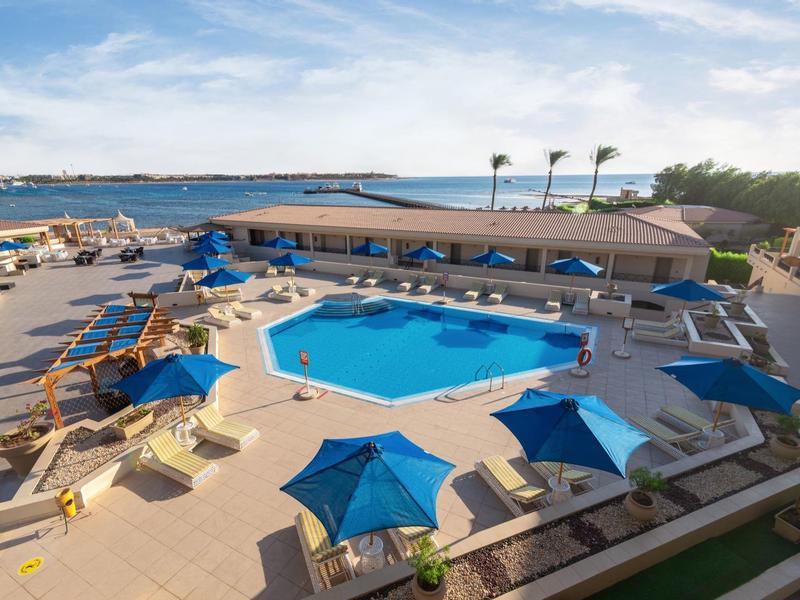 Moderní hotelový bazén s modrými slunečníky a výhledem na moře v pozadí.