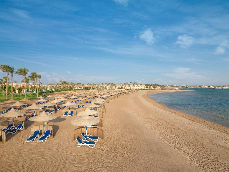 Písčitá pláž s slunečníky a lehátky pod jasnou oblohou podél pobřeží.