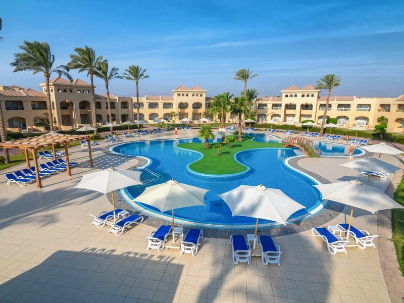 Velký hotelový bazén ve tvaru hvězdy s bílými slunečníky a modrými lehátky, obklopený hotelovými budovami.