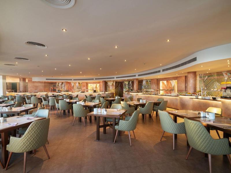 Moderní hotelová restaurace s mnoha stoly a zelenými židlemi, teplé osvětlení a nenápadná výzdoba.