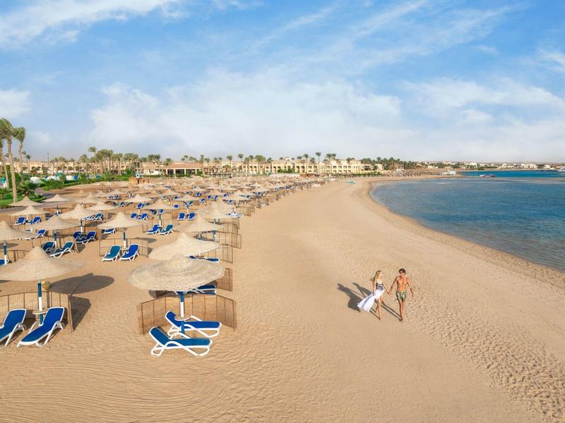 Slunečná písečná pláž s lehátky, slunečníky a několika lidmi u klidného moře.