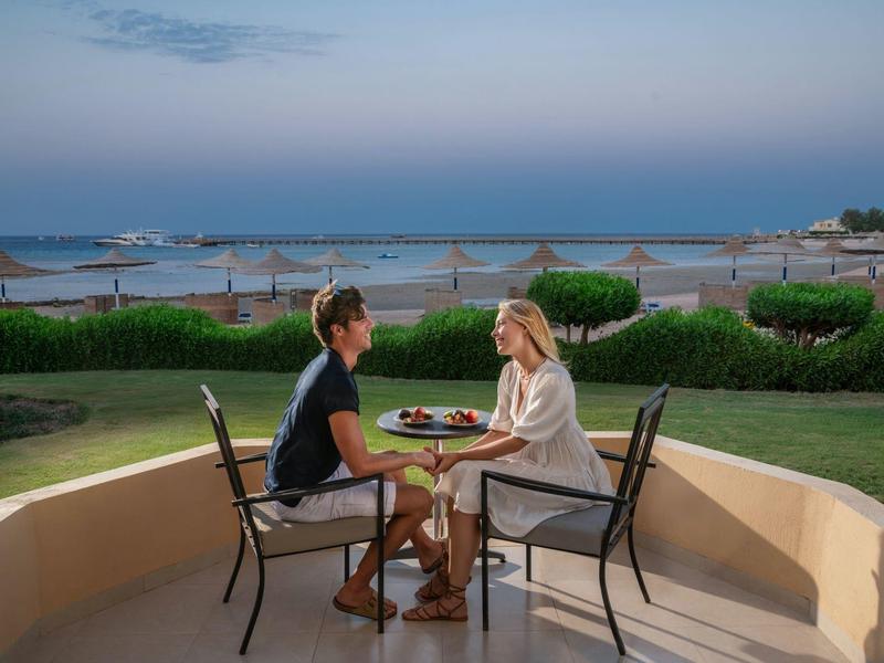 Pár sedí u stolu na terase s výhledem na klidnou pláž a moře při západu slunce.