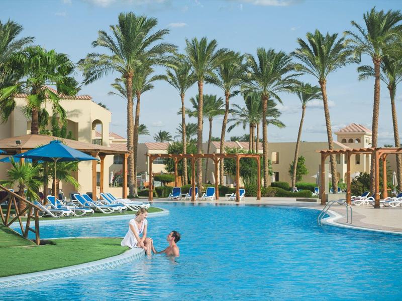 Velký bazén s palmami a lehátky v hotelovém areálu pod modrou oblohou.