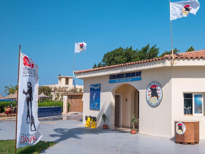 Malá bílá budova s taškovou střechou, vlajkami a jasnou modrou oblohou, obklopená stromy.