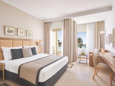 Helles Hotelzimmer mit großem Bett, Holzmöbeln und Blick auf Balkon mit Meer und Palmen.