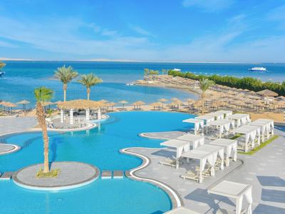 Großer Resort-Pool mit Palmen, weißen Liegen und blauem Meer im Hintergrund unter klarem Himmel.