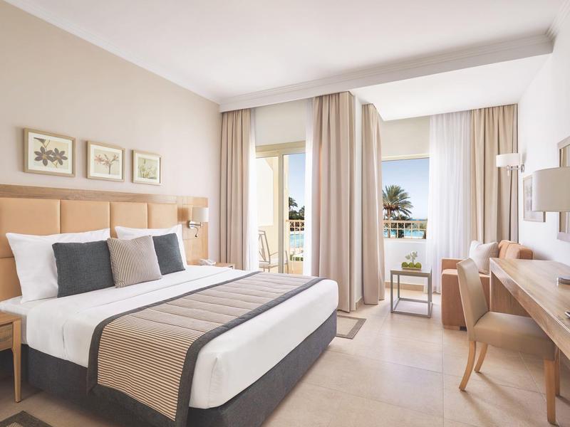 Bright hotel room with a double bed, beige curtains, and a balcony overlooking palm trees and water.