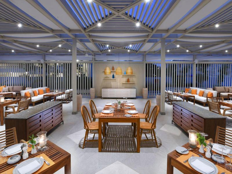 Spacious hotel restaurant with wooden tables, chairs, and modern lighting under a glass ceiling.