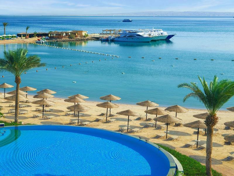 Coastal resort with blue pool, palm trees, sandy beach, umbrellas, and boats in calm sea.
