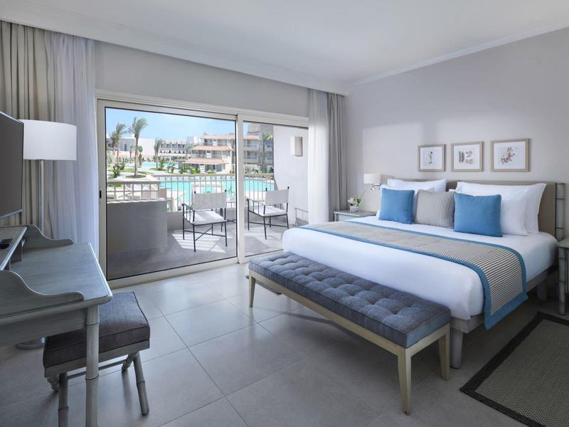 Bright hotel room with king bed, blue cushions, desk, and balcony overlooking pool.