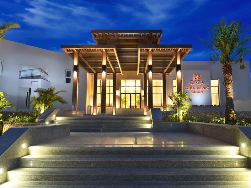 Well-lit hotel entrance with stairs, wooden roof, and palm trees under a twilight sky.