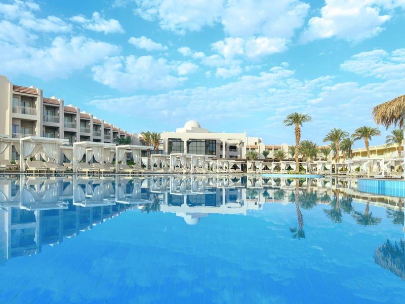 Resort with pool reflecting blue sky, surrounded by lounge chairs and palm trees.