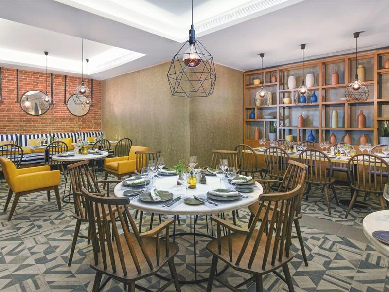 Modern restaurant with wooden chairs, yellow armchairs, round tables set for dining, and decorative ceiling lights.