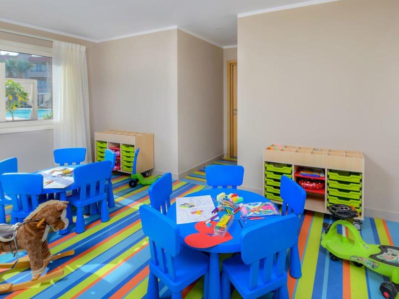 Colorful kids' playroom with blue chairs, tables, toys, and a striped rug by a window.