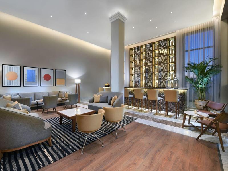 Modern hotel lounge with comfortable seating, striped rug, and a backlit bar with stools.