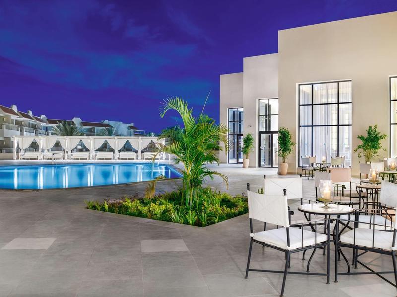 Modern hotel patio with pool, white chairs, tables, and warm evening lighting.