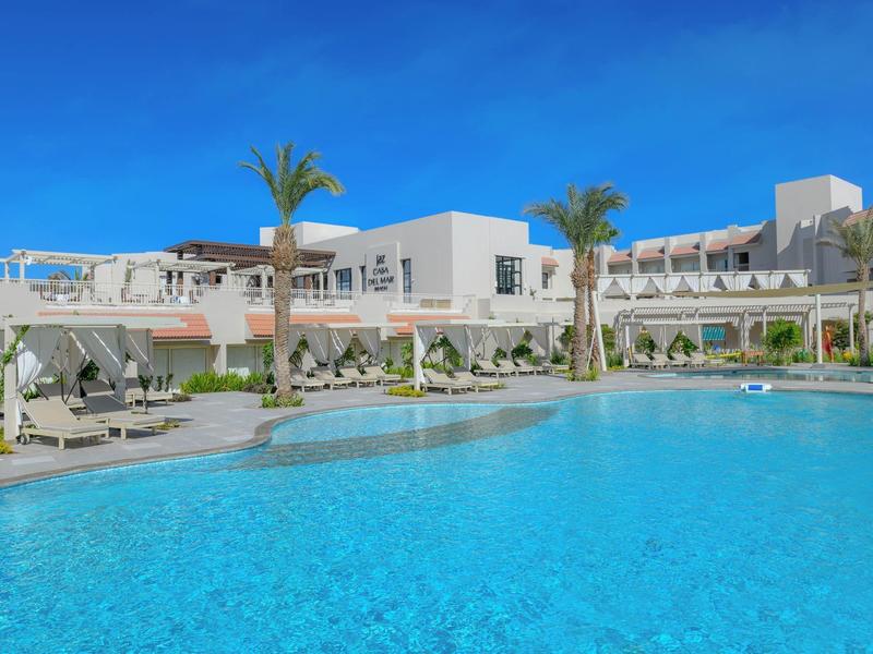 Resort with large pool, sunbeds, and modern white buildings under a clear blue sky.