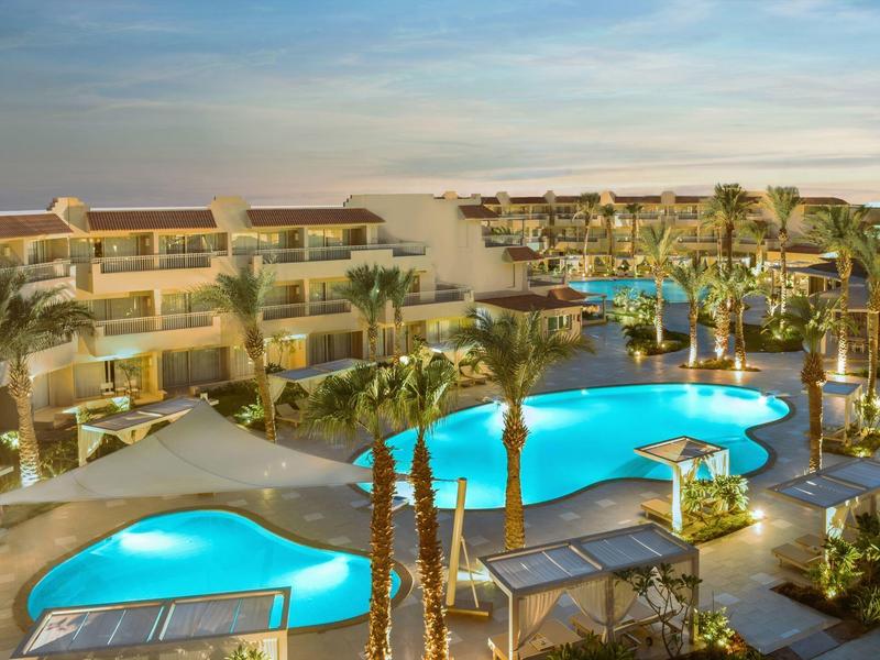 Resort with curvy pools, palm trees, loungers, and beige multi-story buildings under a blue sky.