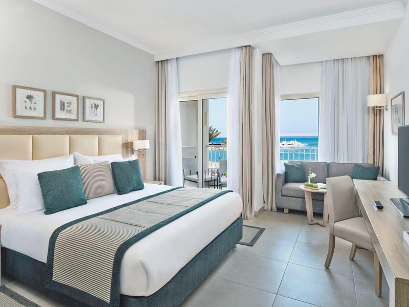 Modern hotel room with king bed, seating area, desk, and sea view through large windows.