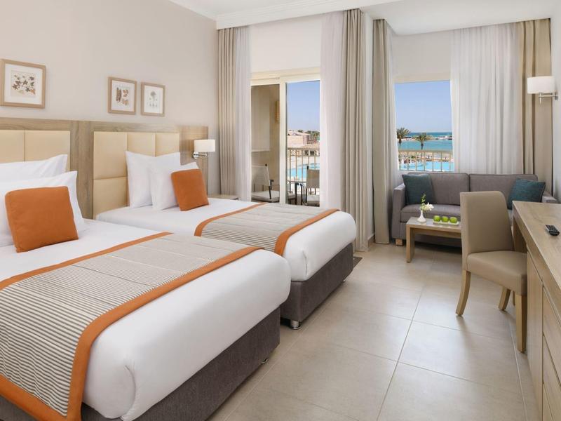 Modern hotel room with two beds, orange accents, balcony, TV, and workspace with a chair.