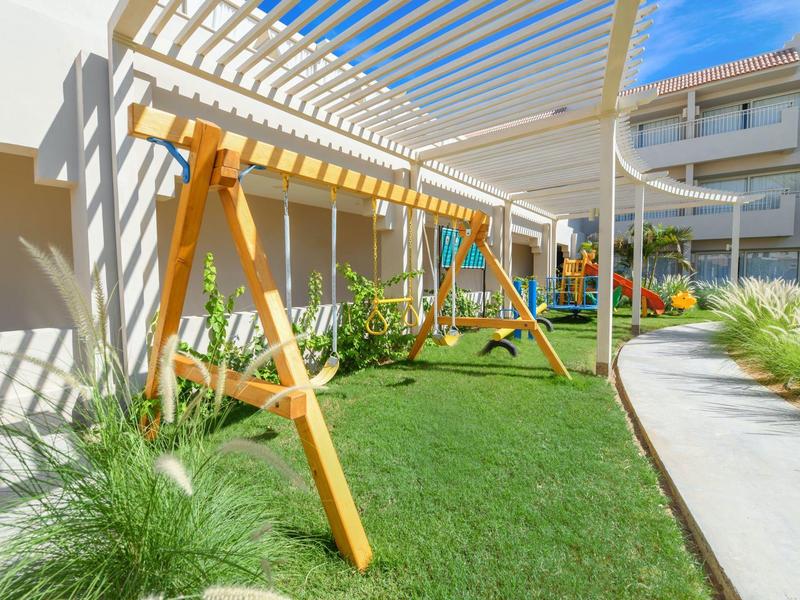 Outdoor playground with wooden swings on a green lawn beside a hotel building under a pergola.