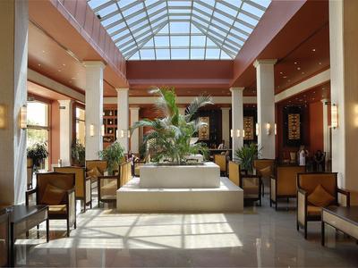 Helle Hotellobby mit zentraler Pflanzeninstallation, Sofas und Stühlen unter einem Glasdach.