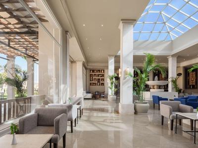 Lichte hotel-loungesectie met modern meubilair, glazen dak, planten en comfortabele zitplaatsen.