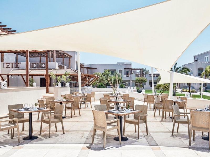 Hotelová terasa s dřevěnými stolky pod bílým slunečníkem, moderní budovy v pozadí.