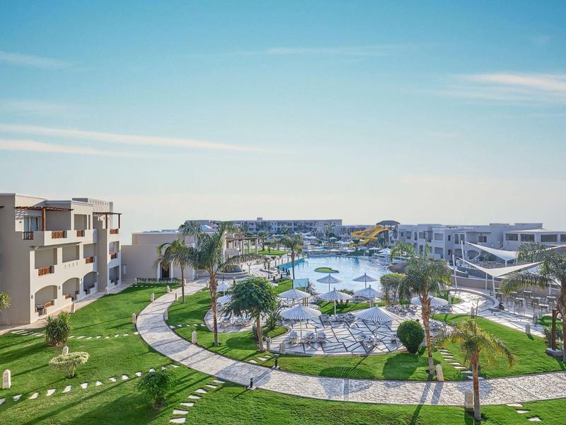 Hotelový komplex s bazénem, palmami a zahradami pod jasnou modrou oblohou.