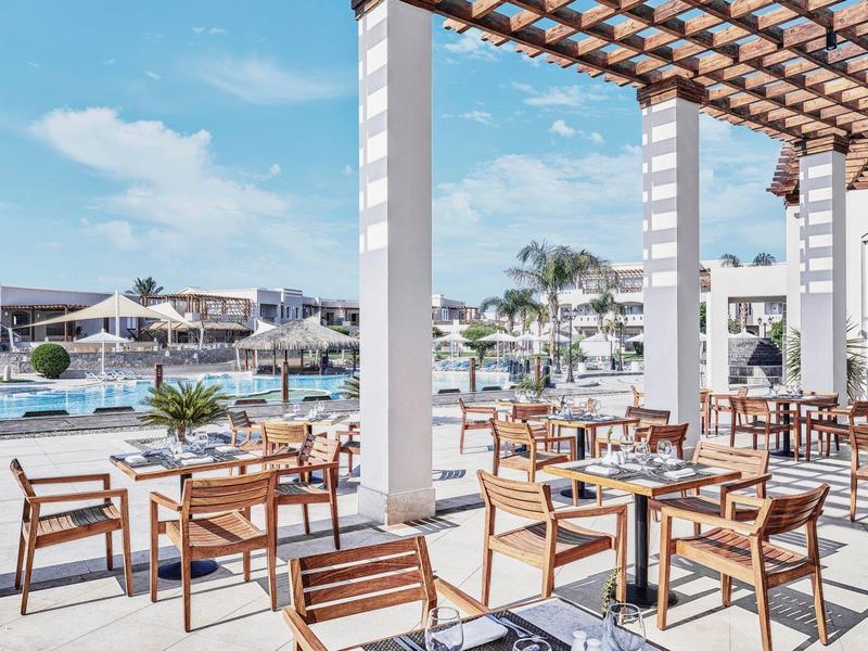 Dřevěná terasa restaurace s výhledem na hotelový bazén pod modrou oblohou.