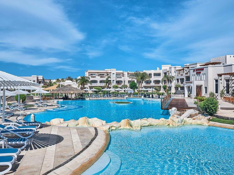 Hotelový bazén obklopený lehátky pod jasnou modrou oblohou.