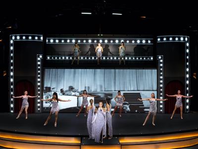 Bühne mit Tänzern und Sängern bei einer Theateraufführung vor einer beleuchteten Kulisse.