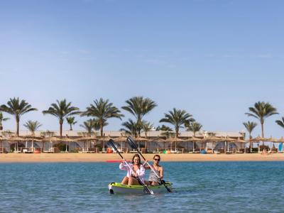 Zwei Personen paddeln in einem Kajak auf ruhigem Wasser vor einem Strand mit Palmen und Sonnenschirmen.