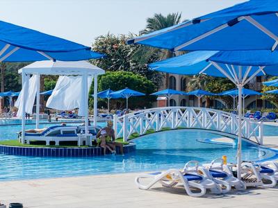 Poolbereich mit blauen Sonnenschirmen, Sonnenliegen und einer kleinen Brücke über das Wasser.